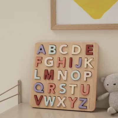 Wooden alphabet letter set on a shelf with a teddy bear and framed picture in the background.
