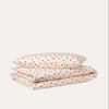 A folded bedding set consisting of a duvet cover and two pillows, featuring a cream-colored fabric printed with a small red heart pattern, set against a solid off-white background.