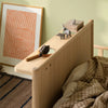 A light natural wood children's bed frame with a built-in shelf, styled with small toy animals and a magnifying glass, set against a pale green wall with a framed abstract print.