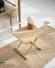 Oliver Furniture Camp bed table in Oak with crossed legs and a small lamp on top, shown beside a canopy bed.