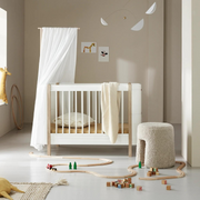 Oliver Furniture Mini+ cot bed in White/Oak beneath a sheer canopy, with a pale blanket draped over the side.