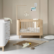 Oliver Furniture Mini+ cot bed in Oak with pale bedding and a mobile above, styled beside a dresser.
