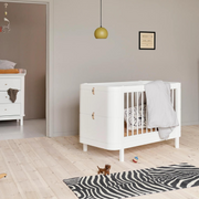 Oliver Furniture white Mini+ cot bed styled with a pale blanket in a nursery with a dresser and zebra-patterned rug.