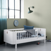 Oliver Furniture white Mini+ junior bed with a short front guard, styled with dark bedding and blue cushions beside a window.