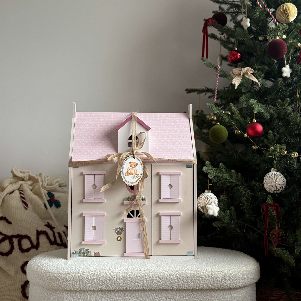 Decorative house-shaped box with a pink roof and white body, placed on a surface with a Christmas tree in the background.