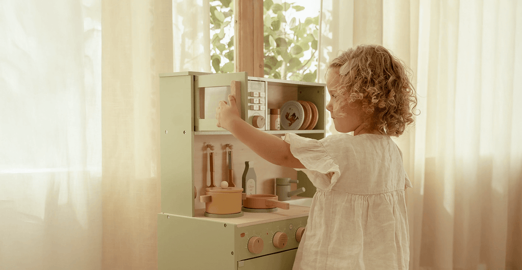 Child playing with a toy kitchen set in a bright room with curtains.