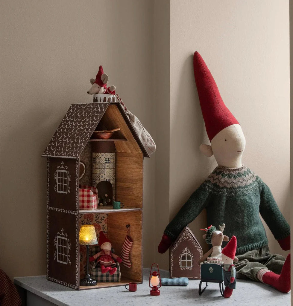 Gnome figurine with a red hat and green sweater next to a wooden house with miniature furniture.