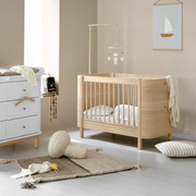Oliver Furniture Mini+ cot bed in Oak with pale bedding and a mobile above, shown at an angle beside a dresser.