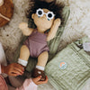 A doll wearing flower-shaped sunglasses lying on an open sage Olli Ella Carrie changing bag as a child adjusts its shoes.