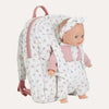 A quilted baby doll backpack with a matching baby doll inside. The backpack is white with a small pink and blue floral print, featuring pink zippers, a pink top handle, and a matching pink bow headband worn by the doll.