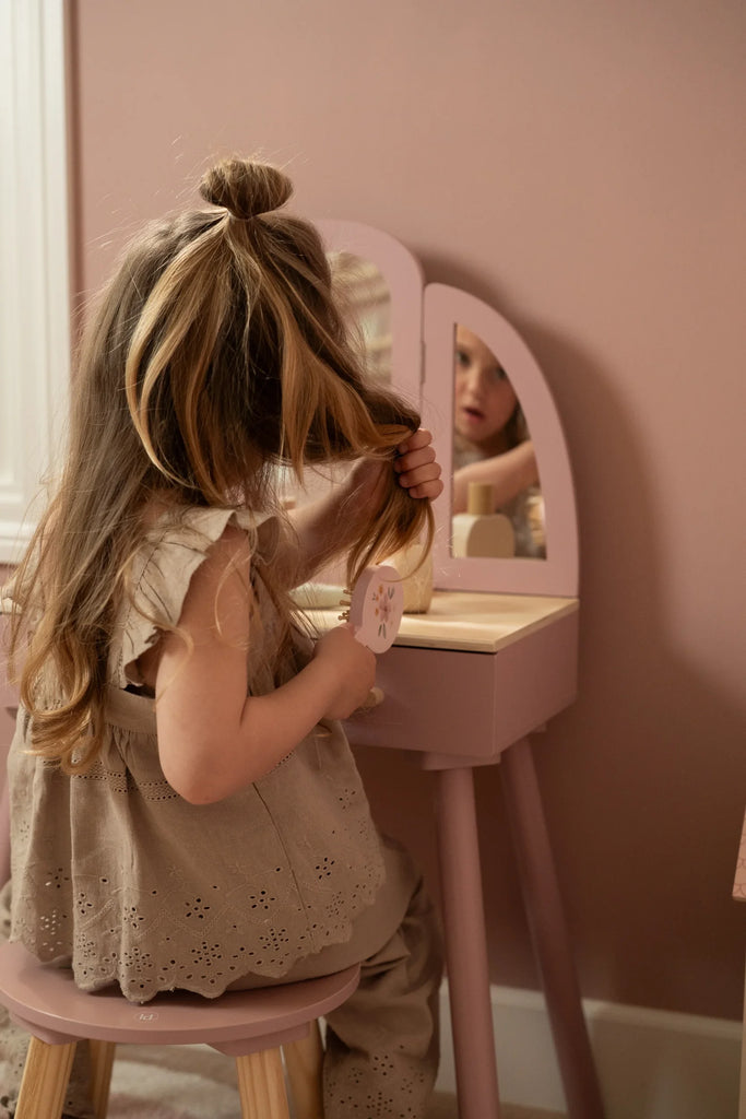 Young girl styling her hair in front of a pink vanity mirror in a softly lit room.