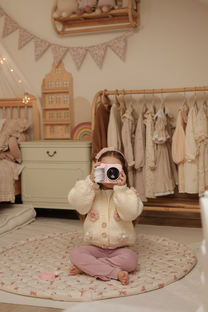 Child in a nursery holding a camera, surrounded by clothes and furniture.