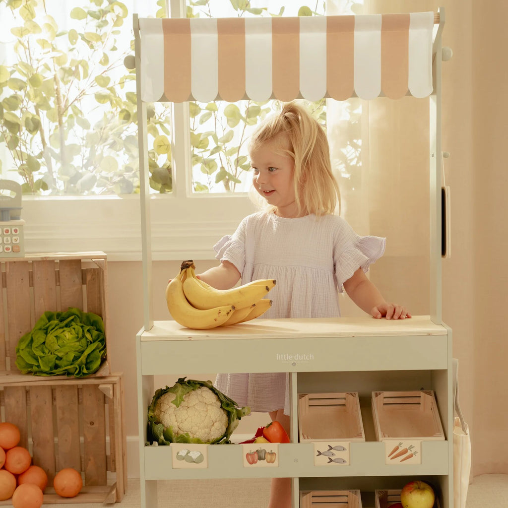 Child playing with a toy kitchen set with fruits and vegetables, standing next to a striped canopy.