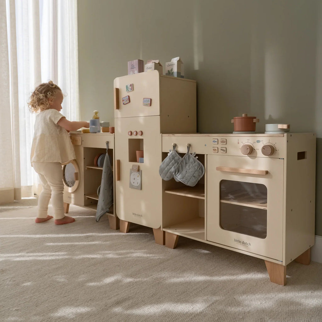 Child playing with a toy kitchen set in a room with large windows.